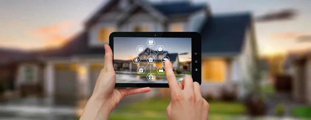 Hände halten Tablet mit Smart Home Steuerungs-App vor modernem Einfamilienhaus