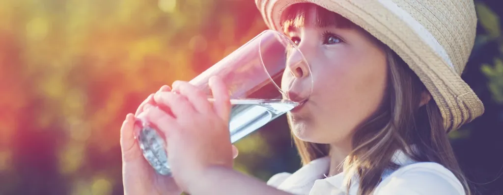 Mädchen mit Strohhut trinkt klares Wasser aus einem Glas im sommerlichen Garten
