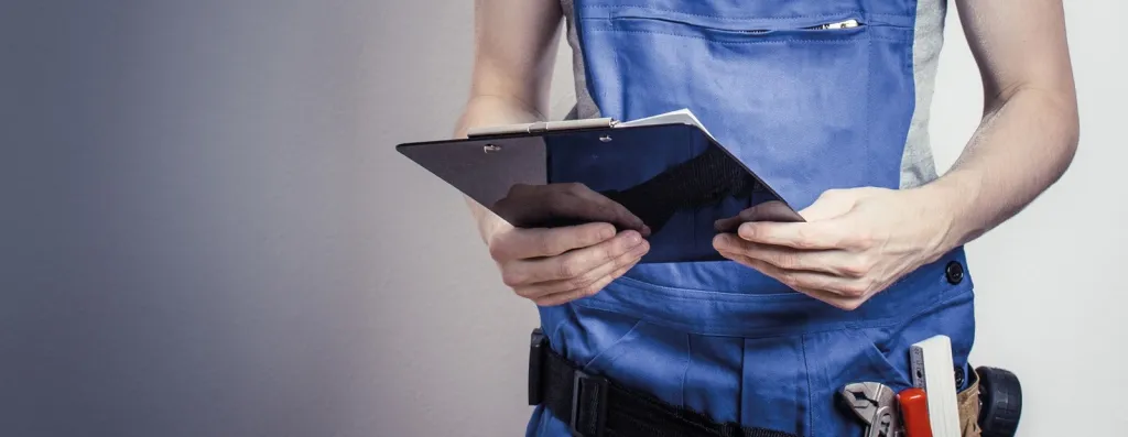 Handwerker in blauer Arbeitskleidung mit Klemmbrett und Werkzeuggürtel bei der Inspektion