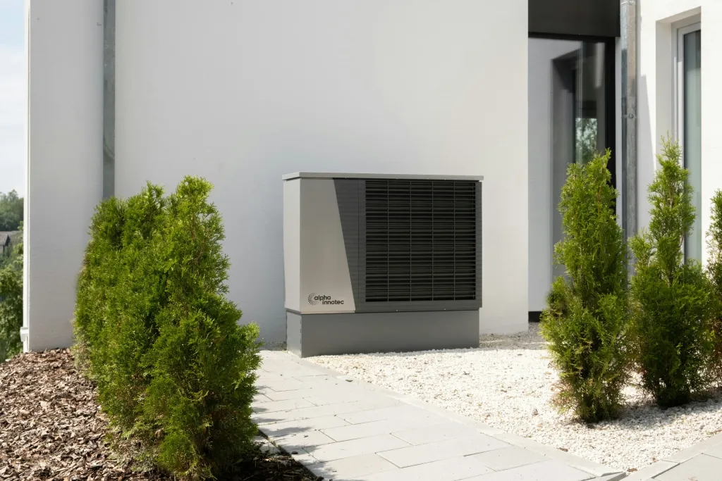 Alpha Innotec Luft-Wärmepumpe an weißer Hauswand zwischen Thujas im Vorgarten